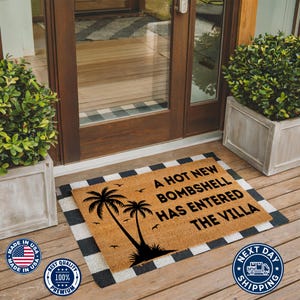 May include: A rectangular coir doormat with a black printed design featuring two palm trees, small birds, and the text A HOT NEW BOMBSHELL HAS ENTERED THE VILLA. The mat is layered over a black and white buffalo check rug on a wooden porch.
