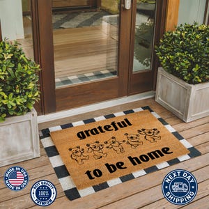 May include: A brown coir doormat with the text "grateful to be home" in black lettering. Five black bear silhouettes are arranged in a line above the text. The doormat is placed on a black and white checkered surface.