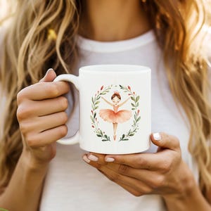 May include: White ceramic mug with a pink ballerina illustration surrounded by green leaves and red berries. The ballerina is wearing a pink tutu and a crown.