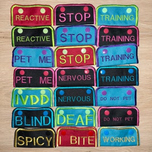 May include: A set of 12 colorful dog training patches with various text, including "Reactive", "Stop", "Training", "Pet Me", "Nervous", "Deaf", "Blind", "Spicy", "Bite", "Working", and "Do Not Pet".