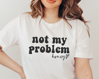 以下が含まれることがあります： 黒いレトロなフォントで「not my problem honey」と書かれた白いTシャツ。茶色の髪と茶色の帽子をかぶった人が着ています。