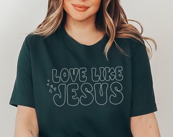 Può includere: Una donna indossa una maglietta verde scuro con il testo "Love Like Jesus" in lettere bianche con contorno. Il testo è in un font a bolle.