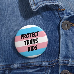 May include: A white button with a blue, pink, and white striped background. The button has the text "PROTECT TRANS KIDS" in black.