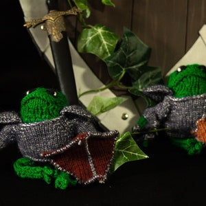 May include: Two green knitted frog plushies wearing gray and brown armor, with one holding a shield. The plushies are sitting on a black surface in front of a wooden shield with ivy.
