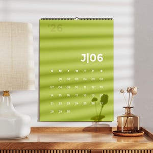Op de afbeelding: Een limoengroene wandkalender voor de maand juni 2026, voorzien van witte tekst en cijfers. De kalender is aan de bovenzijde voorzien van een spiraalbinding en toont de dagen van de week van zondag tot zaterdag, met data van 01 tot 30.