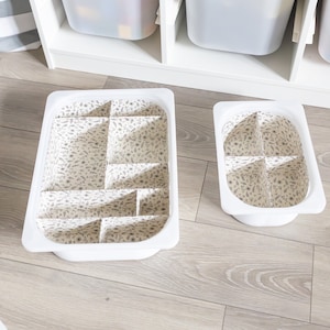 Op de afbeelding: Twee witte plastic opbergbakken met dividers. De bakken zijn bekleed met een stof met een bloemenpatroon. De bakken staan op een lichtbruine houten vloer.