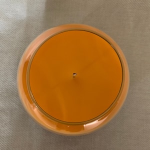 May include: A round orange candle in a clear glass container, viewed from above. The candle has a single wick and a gold-coloured rim. The candle sits on a light grey surface. The candle's diameter is approximately 10 cm.
