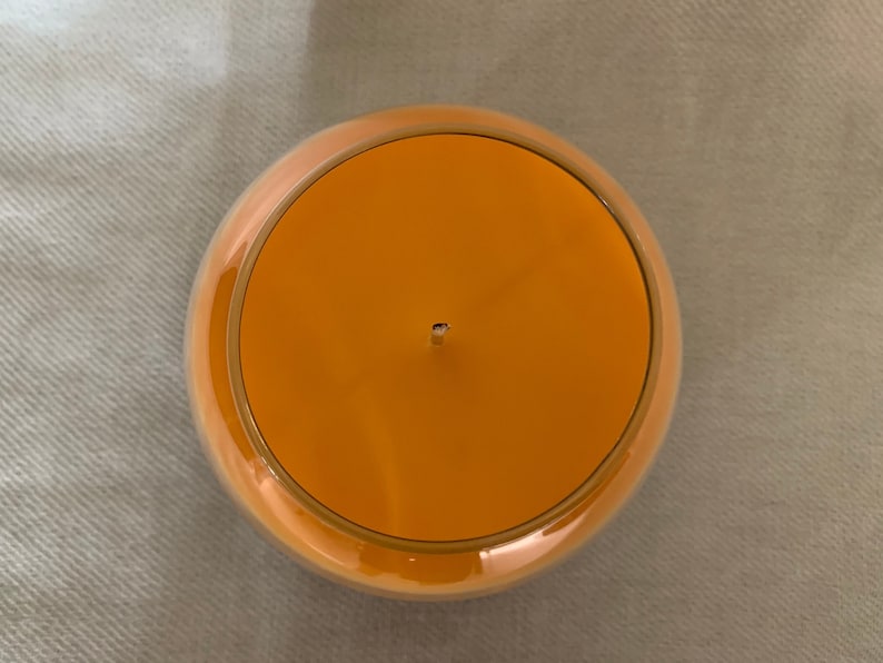 May include: A round orange candle in a clear glass container, viewed from above. The candle has a single wick and a gold-coloured rim. The candle sits on a light grey surface. The candle's diameter is approximately 10 cm.