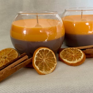 May include: Two spherical glass candles with a layered design. The base is dark brown, topped with orange wax. Dried orange slices and cinnamon sticks are arranged around the candles. Aromatic candles.