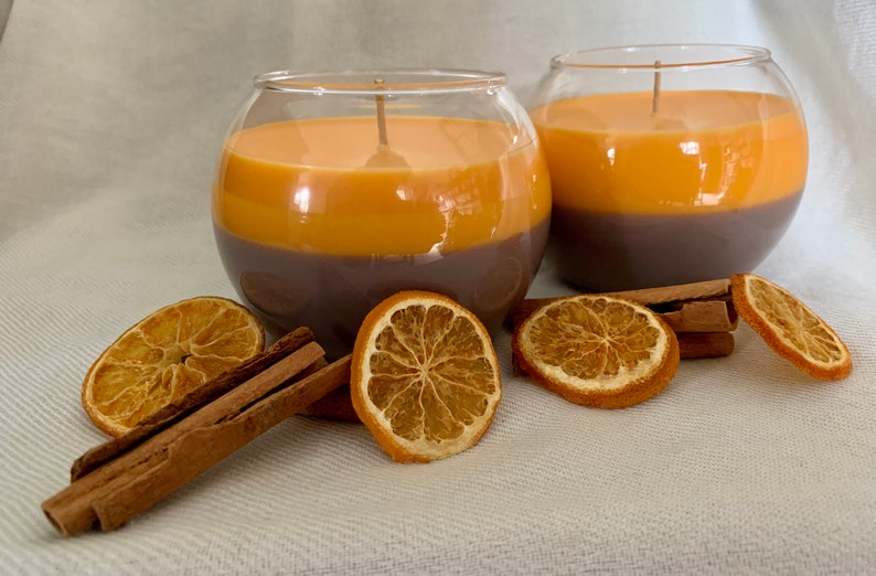 May include: Two spherical glass candles with a layered design. The base is dark brown, topped with orange wax. Dried orange slices and cinnamon sticks are arranged around the candles. Aromatic candles.