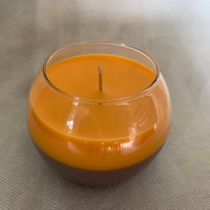 May include: A two-tone candle in a clear glass bowl. The top section is a warm orange, the bottom a deep brown. A single wick is in the centre. The candle sits on a neutral-coloured surface.