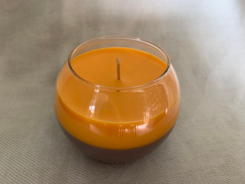 May include: A two-tone candle in a clear glass bowl. The top section is a warm orange, the bottom a deep brown. A single wick is in the centre. The candle sits on a neutral-coloured surface.