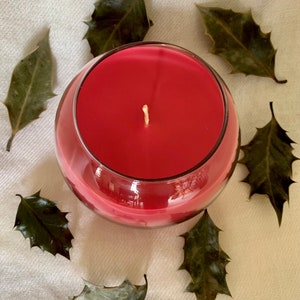 May include: A round, clear glass candle holder filled with red wax. A white wick is centered in the candle. Dried green leaves are arranged around the candle on a cream-colored surface.