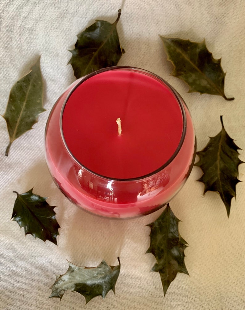 May include: A round, clear glass candle holder filled with red wax. A white wick is centered in the candle. Dried green leaves are arranged around the candle on a cream-colored surface.