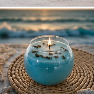 Pode incluir: Uma vela azul acesa em um recipiente de vidro transparente. A vela est&aacute; colocada sobre um tapete redondo e tecido. Pequenos peda&ccedil;os decorativos marrons s&atilde;o vis&iacute;veis na superf&iacute;cie da vela. O fundo mostra uma cena oce&acirc;nica desfocada.