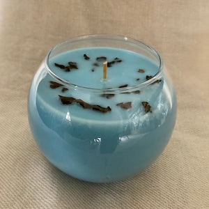 Pode incluir: Um casti&ccedil;al redondo de vidro transparente preenchido com cera azul clara. A vela tem um pavio de madeira e &eacute; decorada com pequenas folhas secas castanhas escuras. A vela est&aacute; sobre uma superf&iacute;cie de cor neutra.
