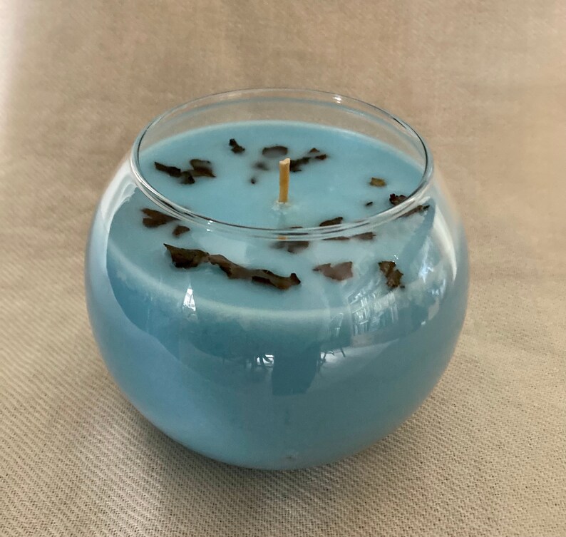 Pode incluir: Um casti&ccedil;al redondo de vidro transparente preenchido com cera azul clara. A vela tem um pavio de madeira e &eacute; decorada com pequenas folhas secas castanhas escuras. A vela est&aacute; sobre uma superf&iacute;cie de cor neutra.