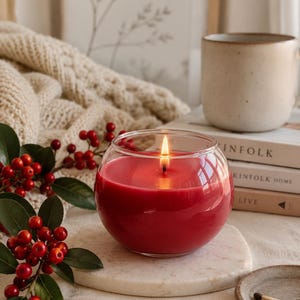 May include: A lit, round, red candle in a clear glass container. The candle sits on a white marble coaster, with a sprig of red berries and a beige mug in the background. A cozy, warm aesthetic.