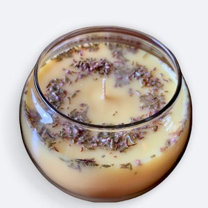 May include: A clear glass jar candle with a yellow wax base and a white wick. The wax is filled with small, dried purple and green botanicals.