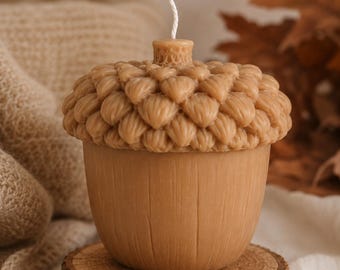 May include: A light brown acorn-shaped candle featuring a textured cap with detailed scale patterns and a smooth, vertically lined base. A single white wick protrudes from the center of the top. The candle is resting on a circular wooden slice.