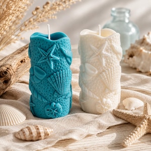 Könnte beinhalten: Zwei zylindrische Kerzen mit Küstenmotiv. Eine ist türkis, die andere weiß. Beide sind mit Seesternen, Muscheln und Seilmotiven verziert. Die Kerzen stehen auf einem beigen Tuch mit Muscheln und einem Seestern.