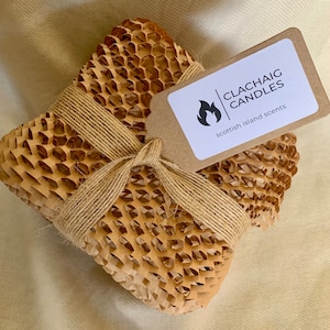 May include: A square package wrapped in honeycomb paper and tied with twine. A small tag with the text "CLACHAIG CANDLES scottish island scents" is attached. The package is on a beige surface.