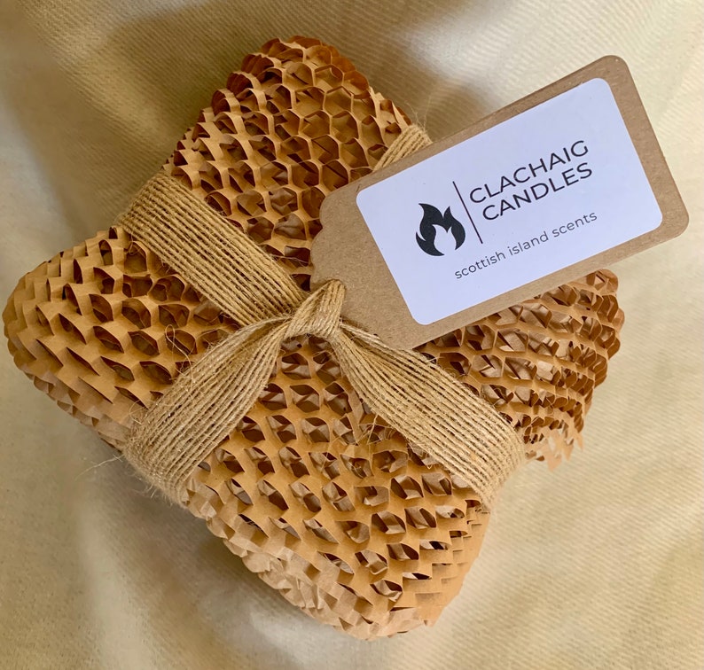 May include: A square package wrapped in honeycomb paper and tied with twine. A small tag with the text "CLACHAIG CANDLES scottish island scents" is attached. The package is on a beige surface.