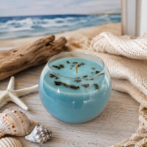 Pode incluir: Um casti&ccedil;al redondo de vidro transparente preenchido com cera azul. A vela tem um pavio de madeira e &eacute; decorada com pequenas folhas verdes escuras. A vela &eacute; rodeada por conchas, uma estrela-do-mar e madeira flutuante, evocando um tema de praia.
