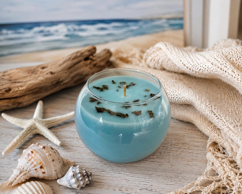 Pode incluir: Um casti&ccedil;al redondo de vidro transparente preenchido com cera azul. A vela tem um pavio de madeira e &eacute; decorada com pequenas folhas verdes escuras. A vela &eacute; rodeada por conchas, uma estrela-do-mar e madeira flutuante, evocando um tema de praia.