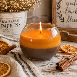May include: A lit, two-tone candle in a clear glass bowl. The candle is orange on top and brown on the bottom. Dried orange slices and cinnamon sticks are arranged around the candle. A small bucket with the text "FARM FRESH PUMPKINS 25&cent;/LB" is in the background.