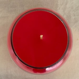 May include: Overhead view of a red candle in a clear glass container. The candle has a smooth, even surface with a single wick centered in the middle. The glass has a rounded shape with a dark rim.