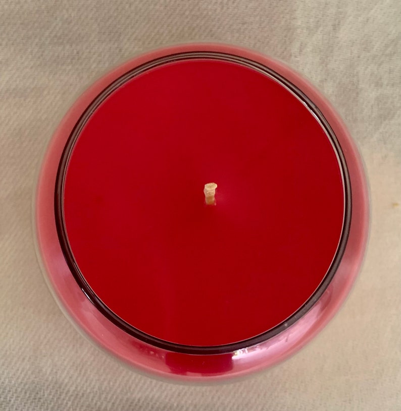 May include: Overhead view of a red candle in a clear glass container. The candle has a smooth, even surface with a single wick centered in the middle. The glass has a rounded shape with a dark rim.