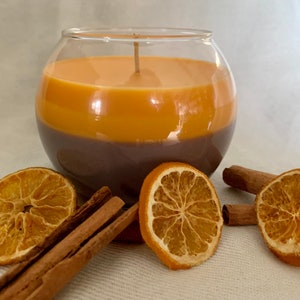 May include: A two-tone candle in a clear glass bowl. The candle is brown at the bottom and orange at the top. Dried orange slices and cinnamon sticks are arranged around the candle, creating a warm, inviting display.