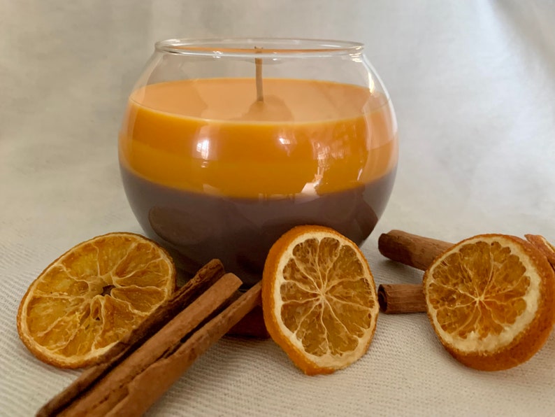 May include: A two-tone candle in a clear glass bowl. The candle is brown at the bottom and orange at the top. Dried orange slices and cinnamon sticks are arranged around the candle, creating a warm, inviting display.