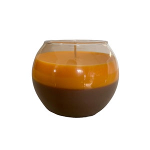 May include: A two-tone candle in a clear glass bowl. The bottom half is a dark brown colour, and the top half is a warm orange. A single wick extends from the centre. The candle is a decorative home accent.