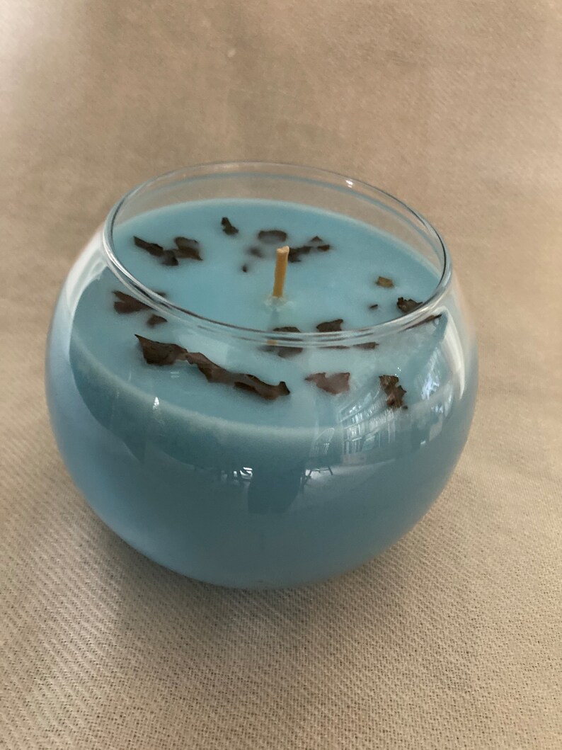 Pode incluir: Um casti&ccedil;al redondo de vidro transparente preenchido com cera azul clara. A vela tem um pavio de madeira e &eacute; decorada com pequenos peda&ccedil;os castanhos escuros. A vela &eacute; um item decorativo.