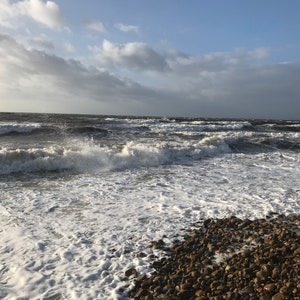 Pode incluir: Uma vista costeira c&eacute;nica com um mar turbulento sob um c&eacute;u nublado. As ondas quebram numa praia de seixos, criando espuma branca. A &aacute;gua &eacute; de um azul escuro e tempestuoso, contrastando com o c&eacute;u mais claro.