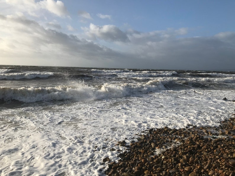 Pode incluir: Uma vista costeira c&eacute;nica com um mar turbulento sob um c&eacute;u nublado. As ondas quebram numa praia de seixos, criando espuma branca. A &aacute;gua &eacute; de um azul escuro e tempestuoso, contrastando com o c&eacute;u mais claro.
