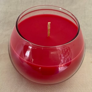 May include: A red candle in a clear, rounded glass container. The candle wax is a solid, vibrant red, and a single, unlit wick is centered. The glass reflects light, creating highlights and shadows.