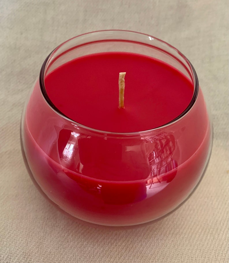 May include: A red candle in a clear, rounded glass container. The candle wax is a solid, vibrant red, and a single, unlit wick is centered. The glass reflects light, creating highlights and shadows.