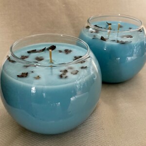 Pode incluir: Duas velas redondas em recipientes de vidro transparente, preenchidas com cera azul clara. A cera &eacute; decorada com pequenas folhas secas castanhas escuras. Cada vela tem um pavio fino e bege.