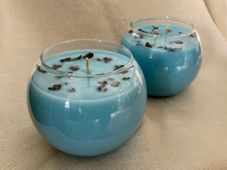 Pode incluir: Duas velas redondas em recipientes de vidro transparente, preenchidas com cera azul clara. A cera &eacute; decorada com pequenas folhas secas castanhas escuras. Cada vela tem um pavio fino e bege.