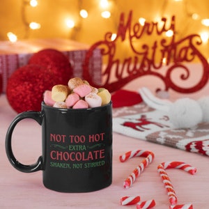 May include: A black mug filled with marshmallows, with the text "NOT TOO HOT EXTRA CHOCOLATE SHAKEN, NOT STIRRED" in red and green. Candy canes and a "Merry Christmas" sign are in the background, suggesting a holiday theme.