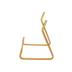 Puede incluir: Un soporte de metal dorado con un diseño simple y minimalista. El soporte está hecho de varillas de metal dobladas, formando una base rectangular y un soporte en ángulo. El soporte está diseñado para sostener un artículo.