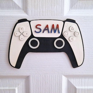 May include: A wooden gaming controller-shaped sign with the name "SAM" in red and blue letters. The controller is black and white, with button details. It is mounted on a white door.
