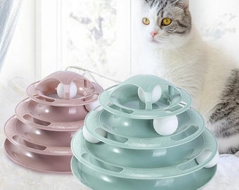 May include: Two cat toy towers in pink and green, each with three tiers and a ball that rolls around the track. A white and gray cat is in the background, looking at the toys.