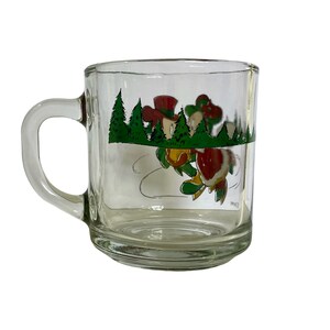May include: A clear glass mug featuring a colorful graphic of a cartoon character wearing a red hat and green clothing, positioned among green pine trees. The design includes a red heart shape and a curved line, with a sturdy handle on the left side.