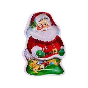 May include: A Santa Claus-shaped serving tray. The tray features a red suit, white beard, and green gloves. A green sack filled with gifts and a teddy bear is visible. A candy cane is also present.