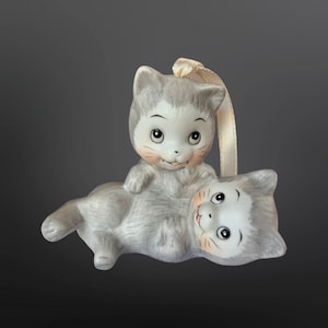 May include: A ceramic ornament featuring two gray kittens playing. The kitten on top has a white face with black eyes and a pink nose. The kitten on the bottom has a white face with black eyes and a pink nose. The ornament has a white ribbon attached to the top.