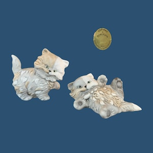 May include: Two porcelain figurines of kittens. One kitten is standing and the other is lying on its back. Both kittens are white and brown with black eyes and pink noses. The figurines are made by Editions Porcelain from Edgerton, Wisconsin.